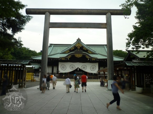 A torii gate and a shrine behind it. The shrine is hanging the imperial banner. The building is made out of dark wood with golden decoration, and the roof is green-grey