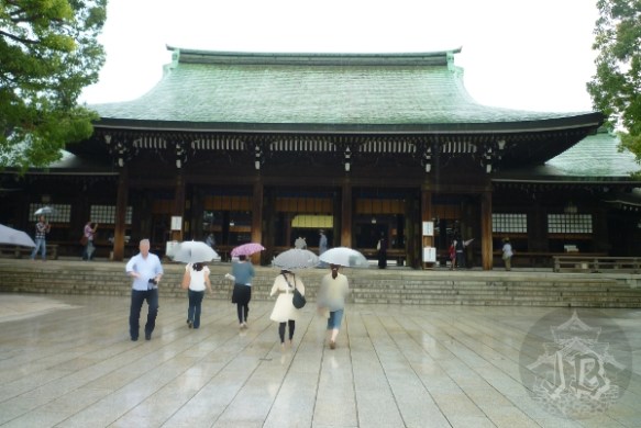 The main building of Meiji Jingu, in dark wood with a grey roof. A few people are approaching the building, all of them are carrying umbrellas
