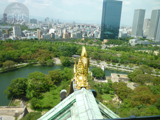 View from the top of Osaka Castle. In the foreground there's the roof decoration, a gold fish-monster. In the background, Osaka highrises. In between the park and the moat