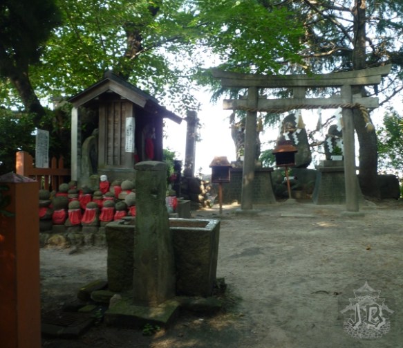Inside a shrine grounds - a little fountain, a stone torii, and a number of small human-like statues wearing red bibs