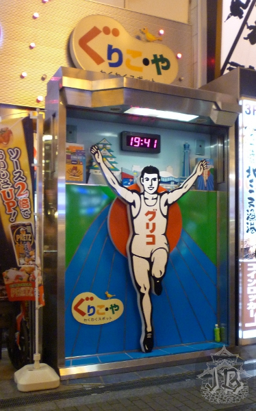 A billboard of a man reaching the end line of an athletic course, raising his arms in victory. His shirt reads グルコ