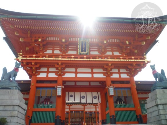 A shrine building, in powerful orange. The sun is shining at the rim of the roof, and there are two sculptures of foxes sitting on the sides.