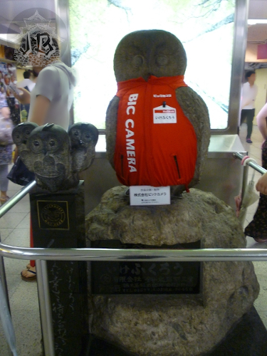 Sculpture of a big barn owl with a red vest. There are three smaller owls on the left, one of them is carrying a heart