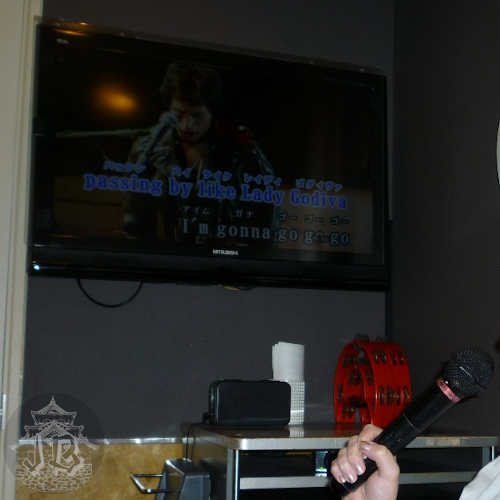 A karaoke screen reading Passing by like Lady Godiva. On the bottom-right corner, there's someone's hand holding a microphone