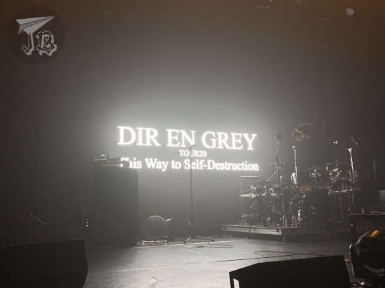 A dark stage with a drum set. Letters projected on the screen behind the stage read Dir en Grey Tour 20 This Way to Self-Destruction