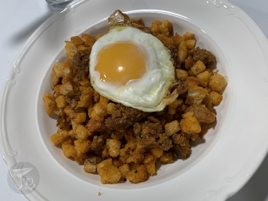 A plate of migas. The breadcrumbs look golden, and there are pieces of pancetta and chorizo mixed with the bread. On top, there is a fried egg, sunny-side up