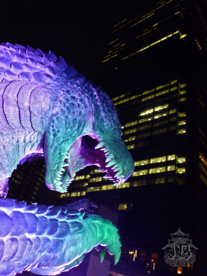 A close-up of the Godzilla statue at night, lit purple and blue, and looming on a skyscraper