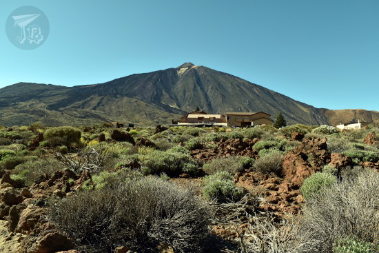 Mount Teide rises in the background. It looks wrinkled due to the different eruptions. At its foot, a low building, looking completely out of place. In the foreground, small bushes in grey and green.