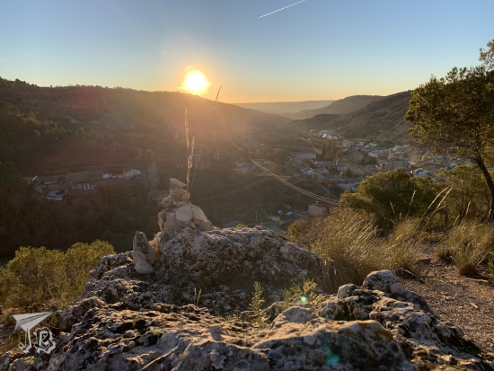Sunset picture. The sun is sinking behind a low mountain. The mountain gives way to a valley where the village is peeking. On the foreground, there is a capricious-looking grey rock. The sun is a big gold ball flaring on everything