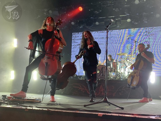 Apocalyptica playing at La Riviera. They have classical cellos. Two of the members stand on the sides, playing their cellos. Another one is walking swinging his as if it weighed nothing, The final one is slamming drums in the background.