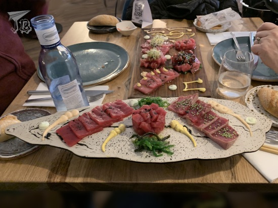 Lunch at DeAtún. A plate with three tuna cuts. The centre is round, and rose-like, and the sides are extended on a line. The fish is uncooked and it looks dark red. There's a similar dish in the background, with more cuts