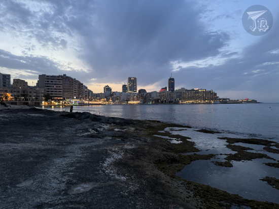Balluta Bay at dusk. The ocean is calm, and the buildings at the other side of the water have started turning on their lights